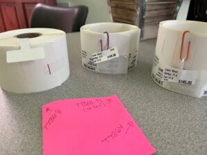 A row of white label rolls with price tags and purple/orange loops on a gray countertop, pink sticky note in foreground.